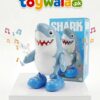 A promotional image of a blue-and-white battery-operated shark toy with a smiling face, cartoon-style teeth, and blue shoes displayed on a white pedestal. The toy is shown in front of its matching retail box labeled "SHARK," with colorful musical notes around it indicating sound or music features. The Toywala.pk logo appears at the top against a clean white background.